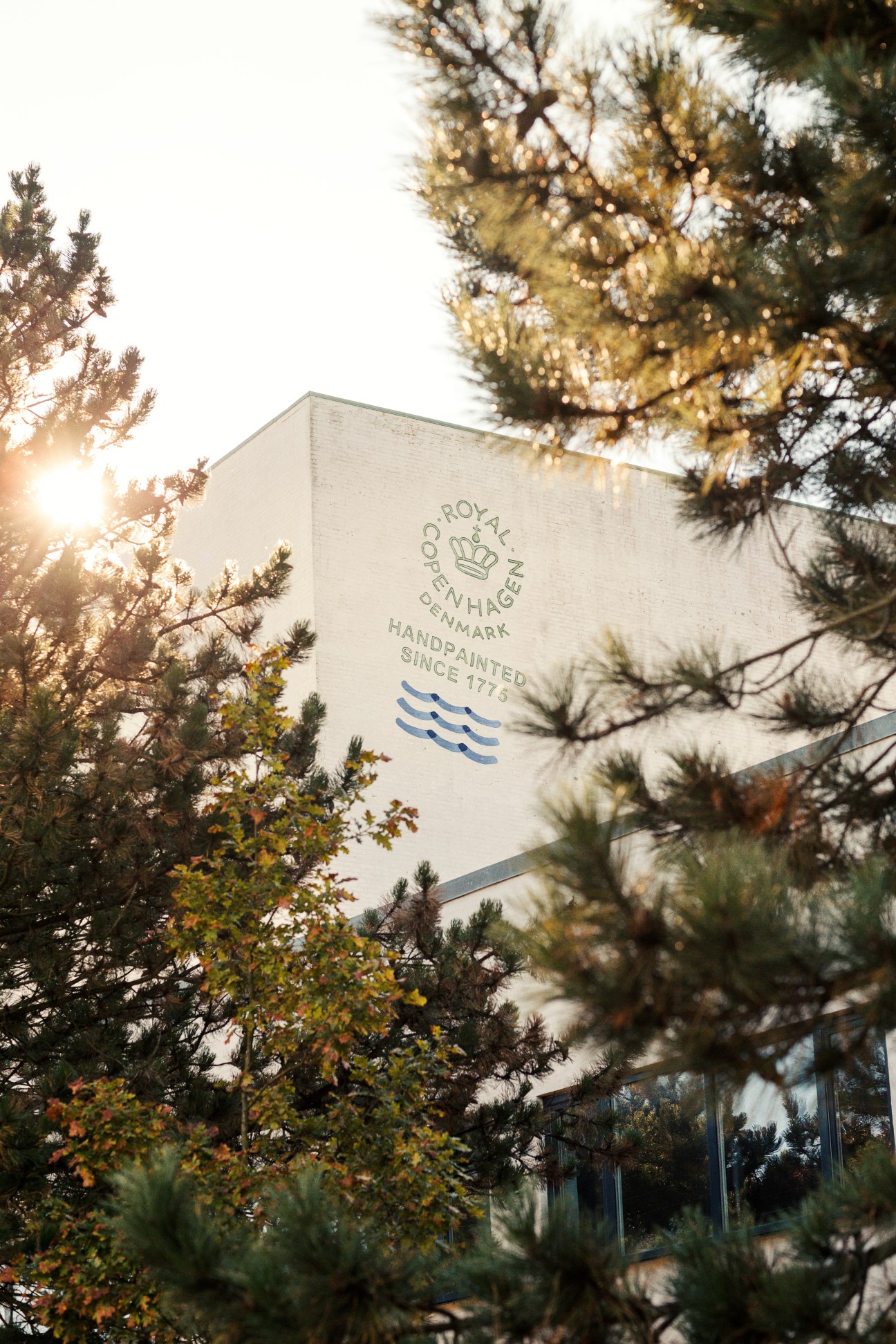 Royal Copenhagen headquarters building with signage visible among evergreen trees at sunset.
