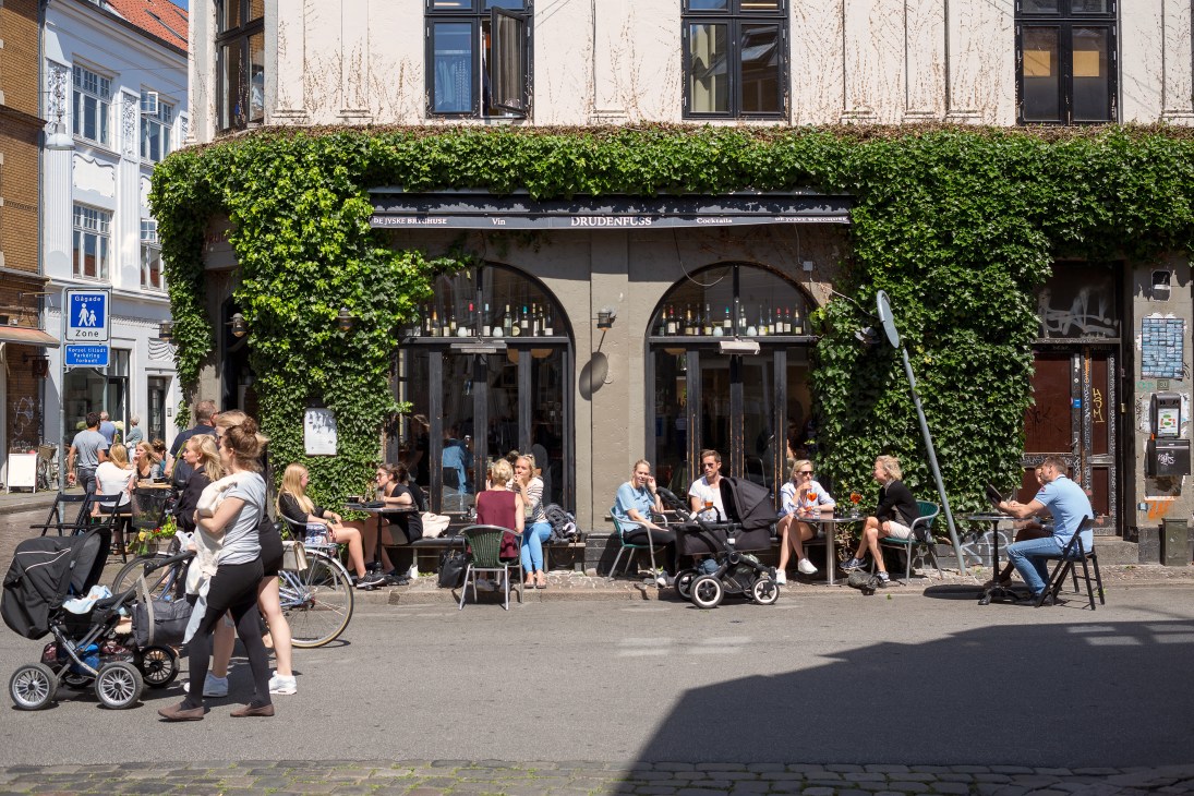 A lively street scene with ivy-covered café storefront and diners seated outdoors on Graven in central Aarhus.
