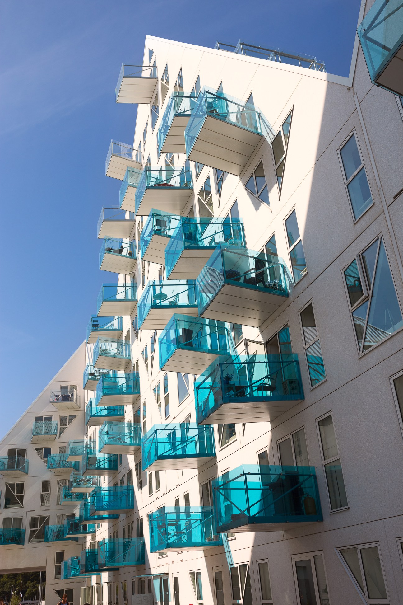 Modern residential building with white facade and staggered turquoise glass balconies against blue sky.