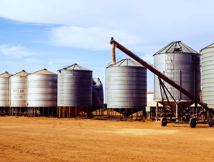 Multiple silver cylindrical grain silos with conical roofs and an auger on a dusty farm yard in Western Australia.