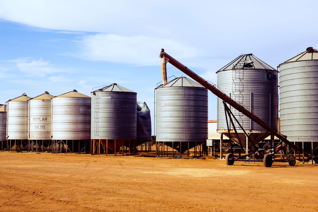 Multiple silver cylindrical grain silos with conical roofs and an auger on a dusty farm yard in Western Australia.