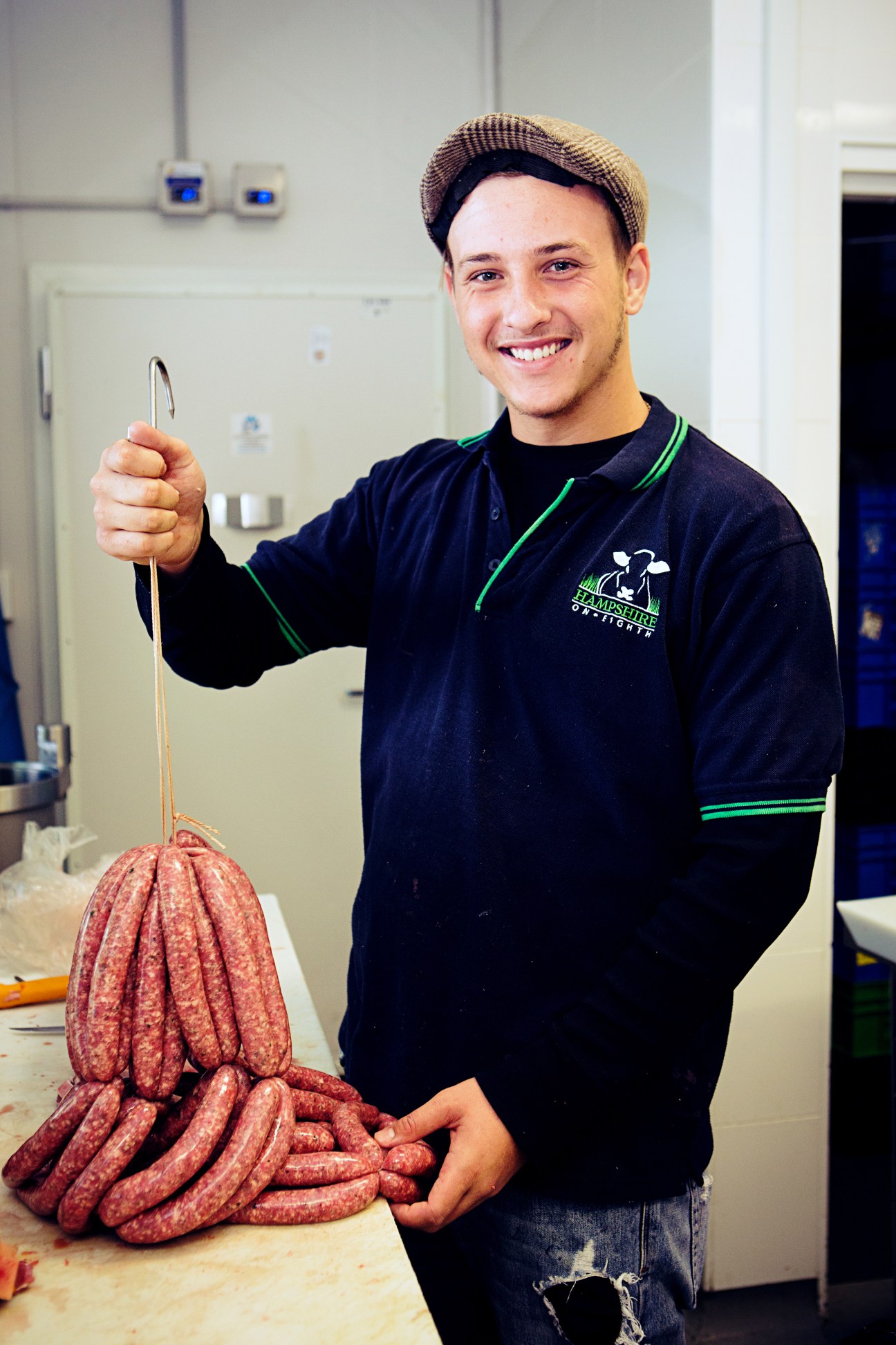 A smiling butcher in a navy apron holds a bundle of red sausages at Hampshire on 8th.