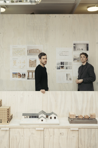 Two men standing behind a white table with architectural models and drawings on wall behind them