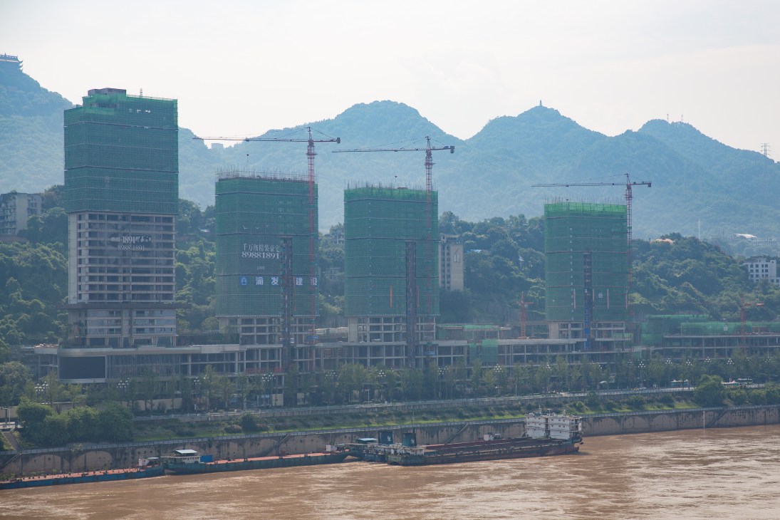 Chongqing skyline with construction cranes, green-wrapped buildings under development, mountains and muddy river in background.