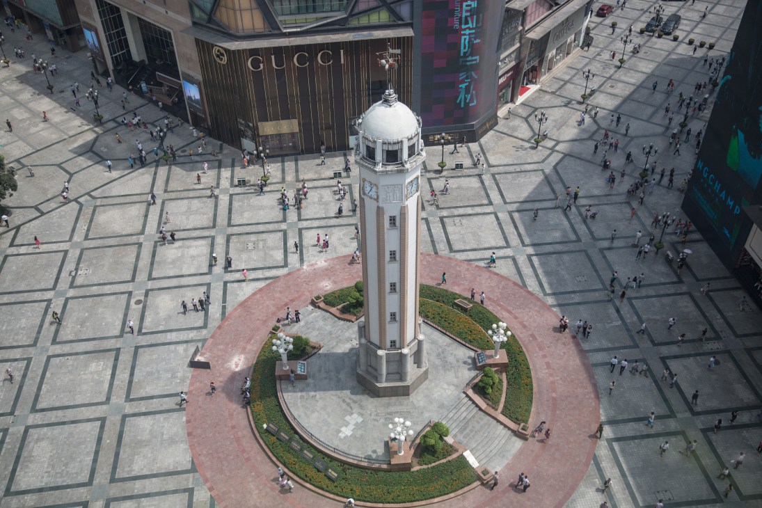Aerial view of the People's Liberation Monument tower surrounded by a plaza with Gucci and other luxury brand storefronts.