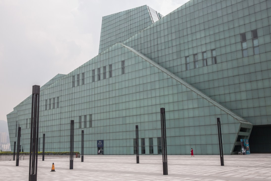 Modern glass and steel theatre building with angular roof design and empty plaza with black bollards