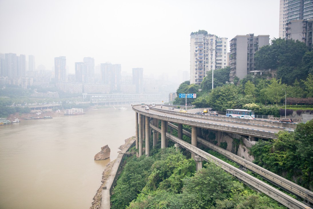 Elevated highway bridge crossing the muddy Jialing River with Chongqing's high-rise buildings and hazy skyline in the background.