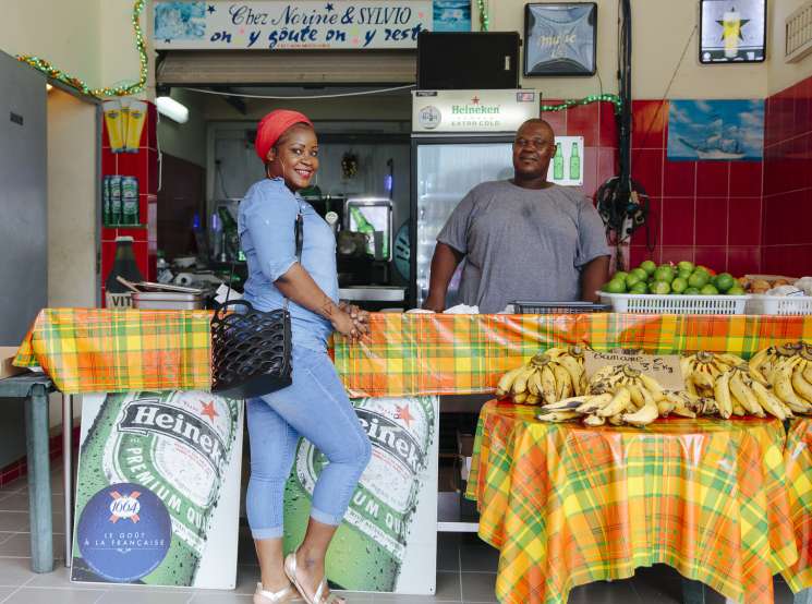 Woman and man standing behind a colorful table at a fruit stand displaying bananas and other produce in Cayenne.