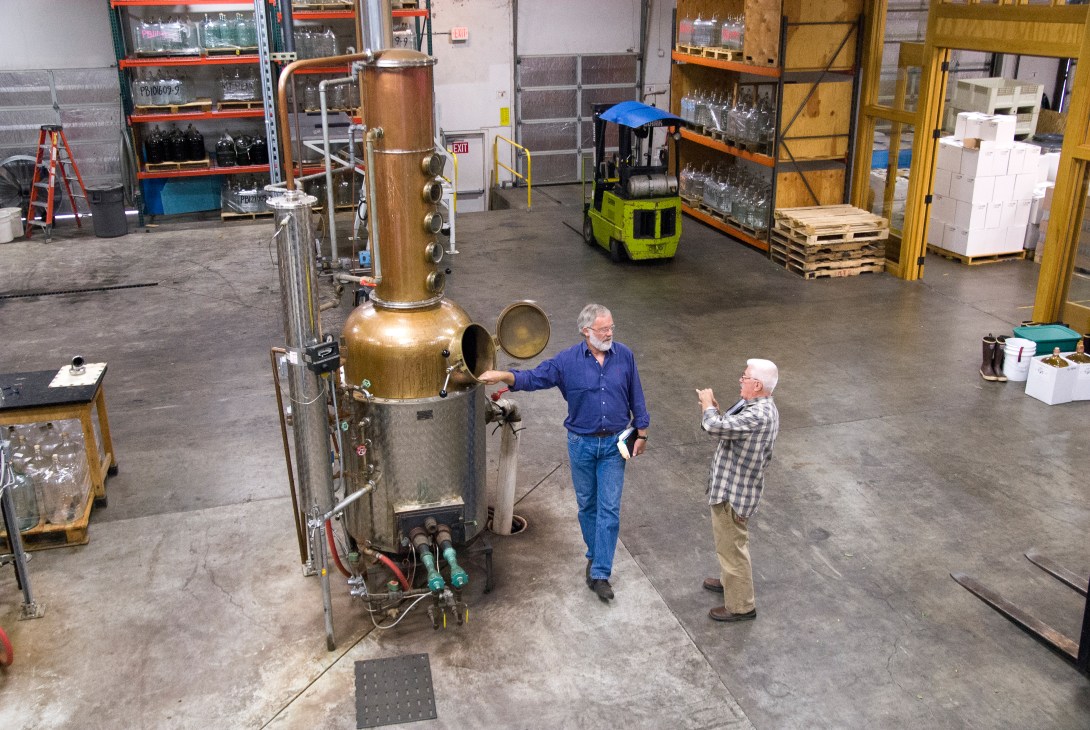 Bill Owens discusses apple brandy next to a copper pot still in a distillery warehouse.