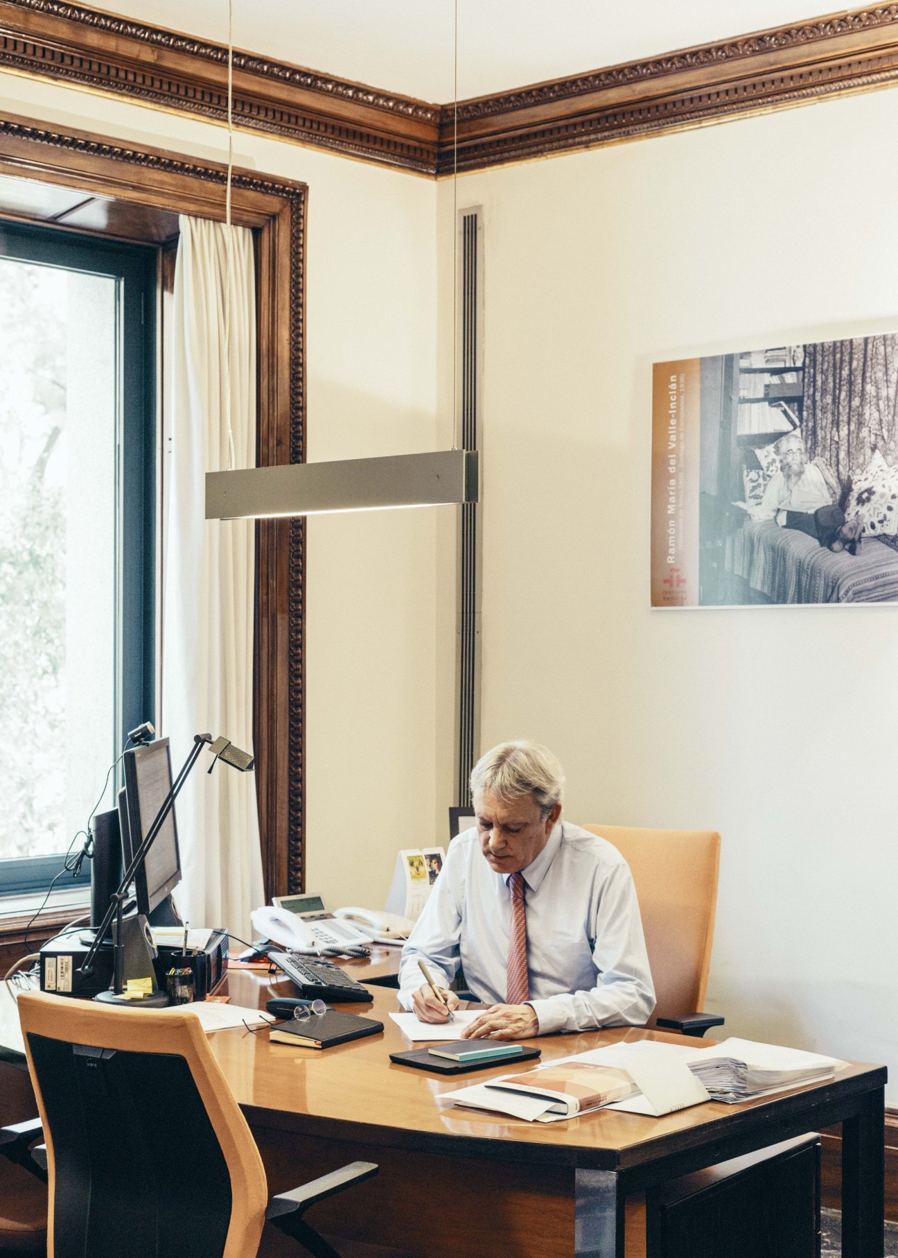 Man in white shirt and tie writing at desk in ornate office with wooden panelling