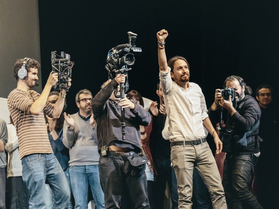 Pablo Iglesias raises his fist in celebration surrounded by camera operators and media crew at a Podemos rally.