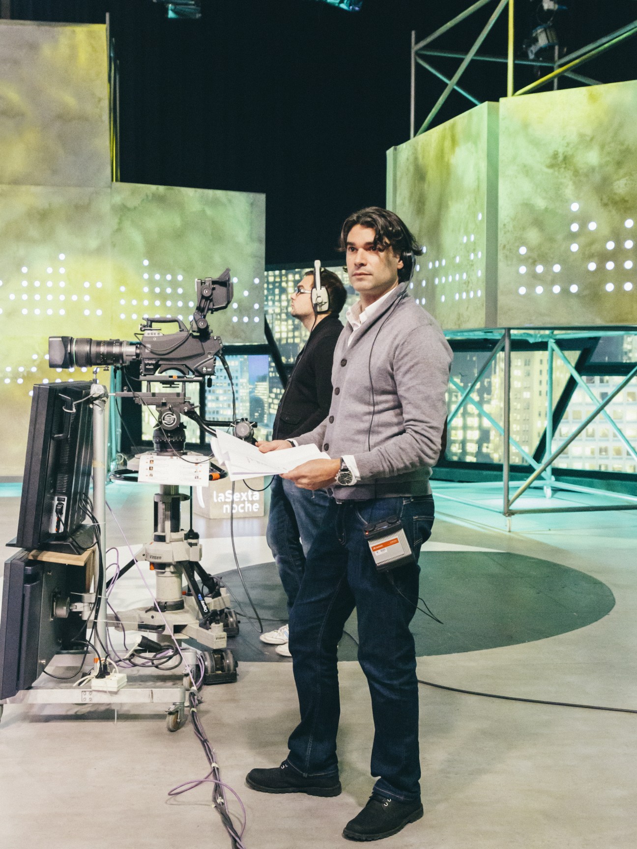 A man in a gray cardigan holds papers while standing next to a television camera on a studio set.