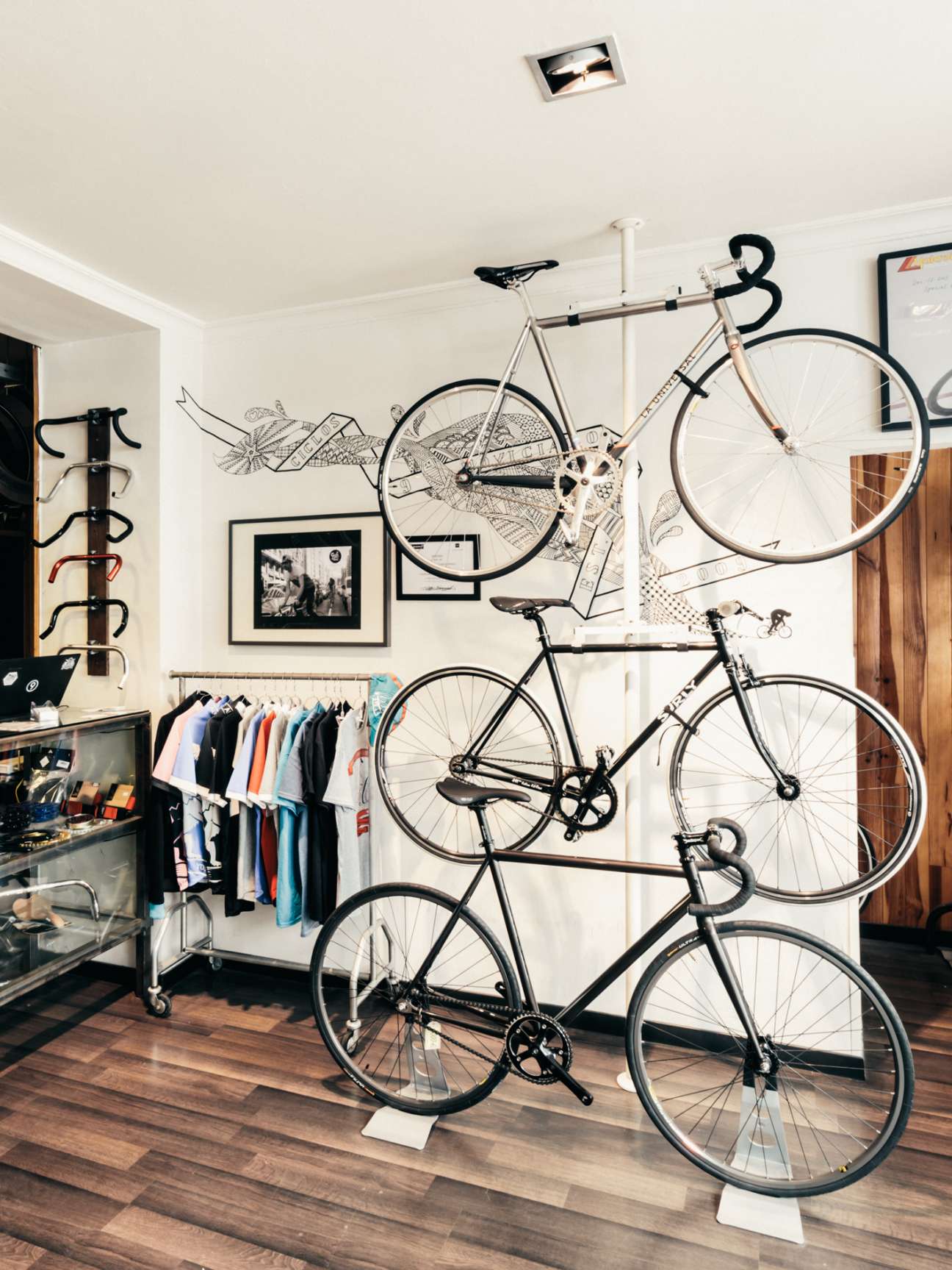 Bicycles mounted on white wall of Ciclos La Universal shop with clothing rack and framed artwork.