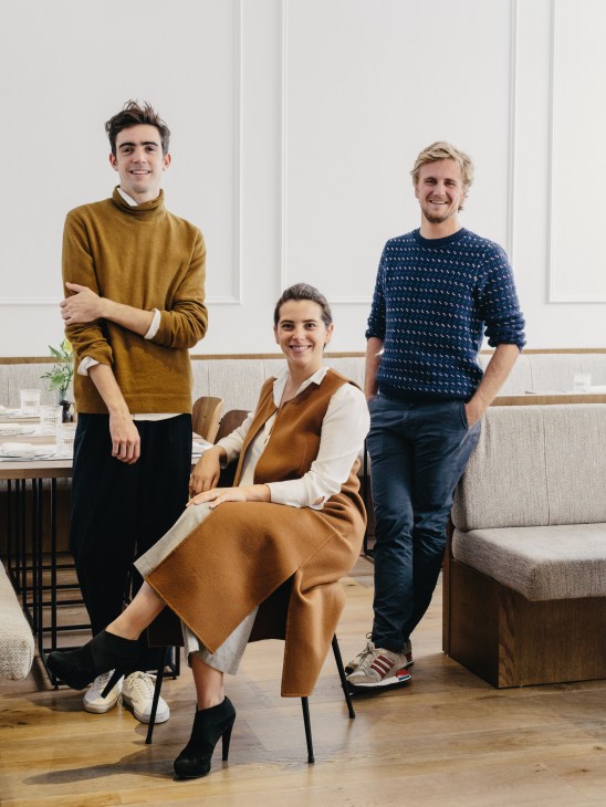 Three people posing in a modern dining space: a man in a mustard sweater, a woman in a brown vest seated, and a man in a navy patterned sweater.