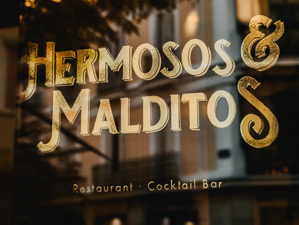Gold lettering reading 'Hermosos & Malditos' with 'Restaurant · Cocktail Bar' underneath on a window or storefront.