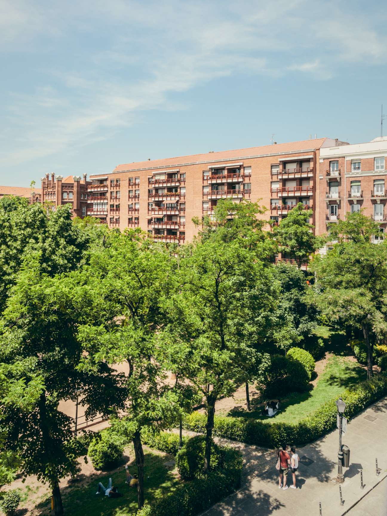 Red-brick residential building overlooking a tree-filled plaza in Madrid's Conde Duque neighbourhood.