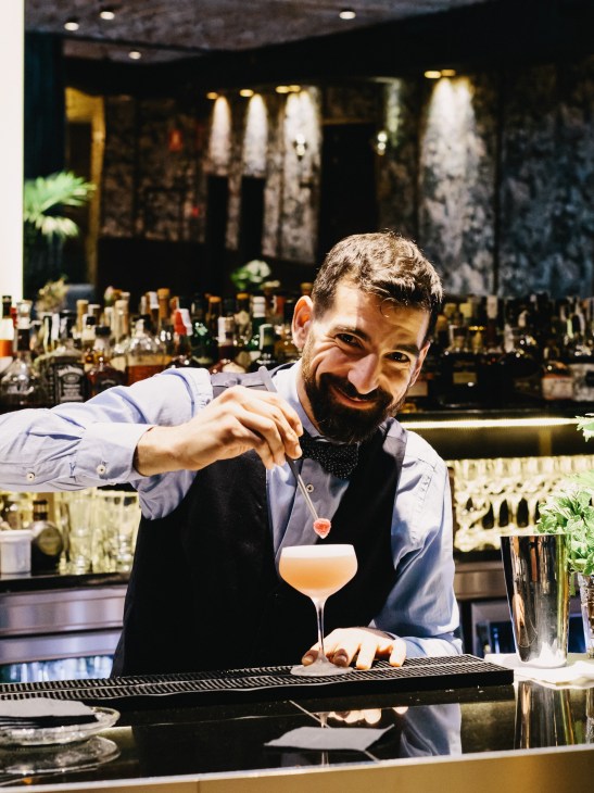 Bartender in blue shirt and black vest preparing a cocktail at a bar with bottles in background.