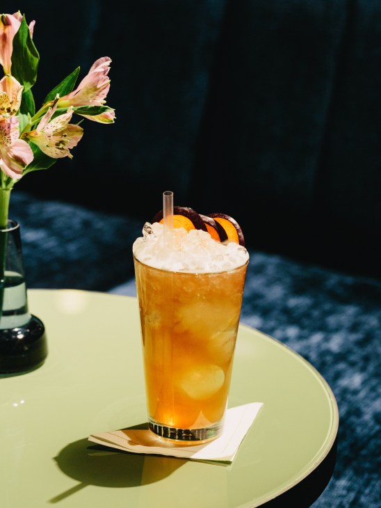 An amber-colored cocktail with ice and whipped cream, garnished with an orange slice and striped cookie, beside pink flowers.