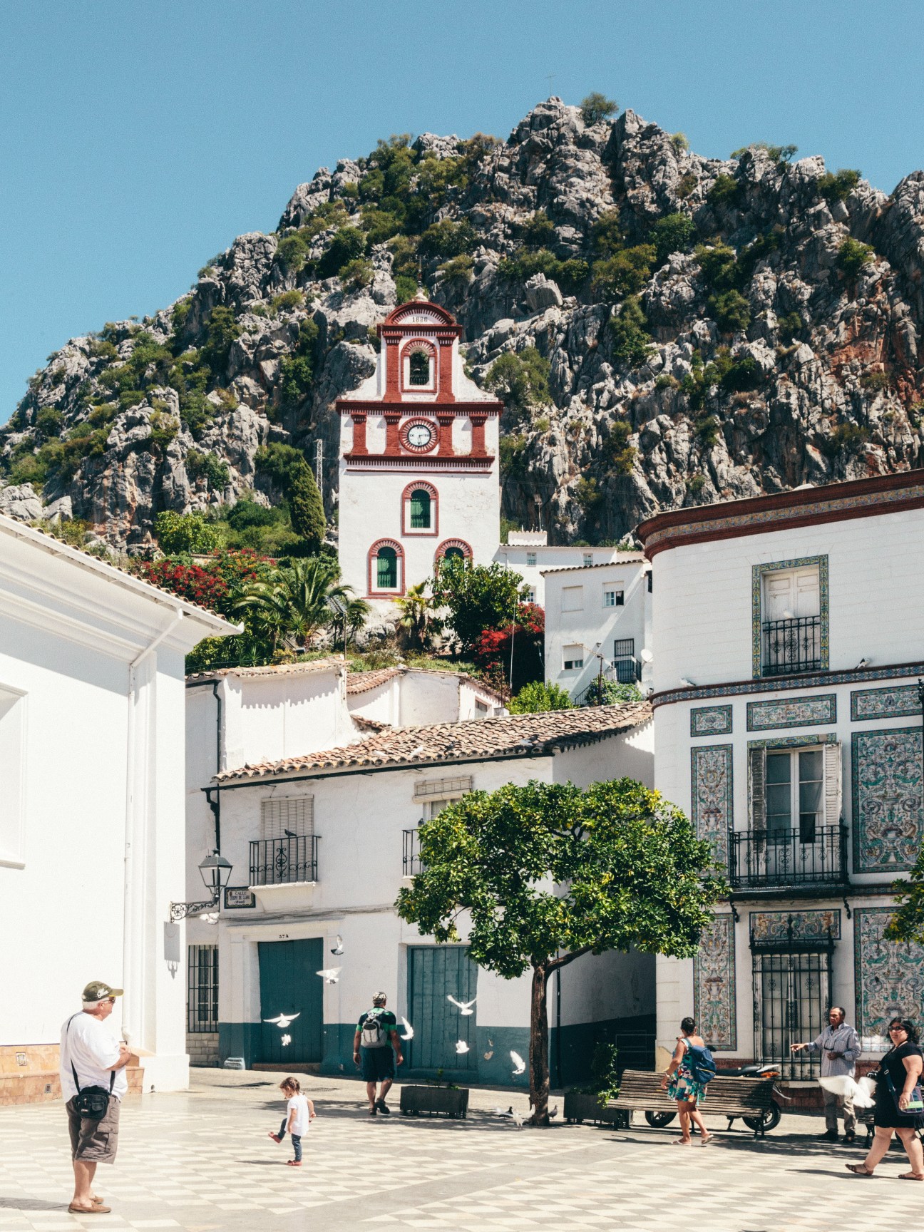 Ubrique town plaza with white-washed buildings, a red and white chapel tower, and limestone mountain backdrop.