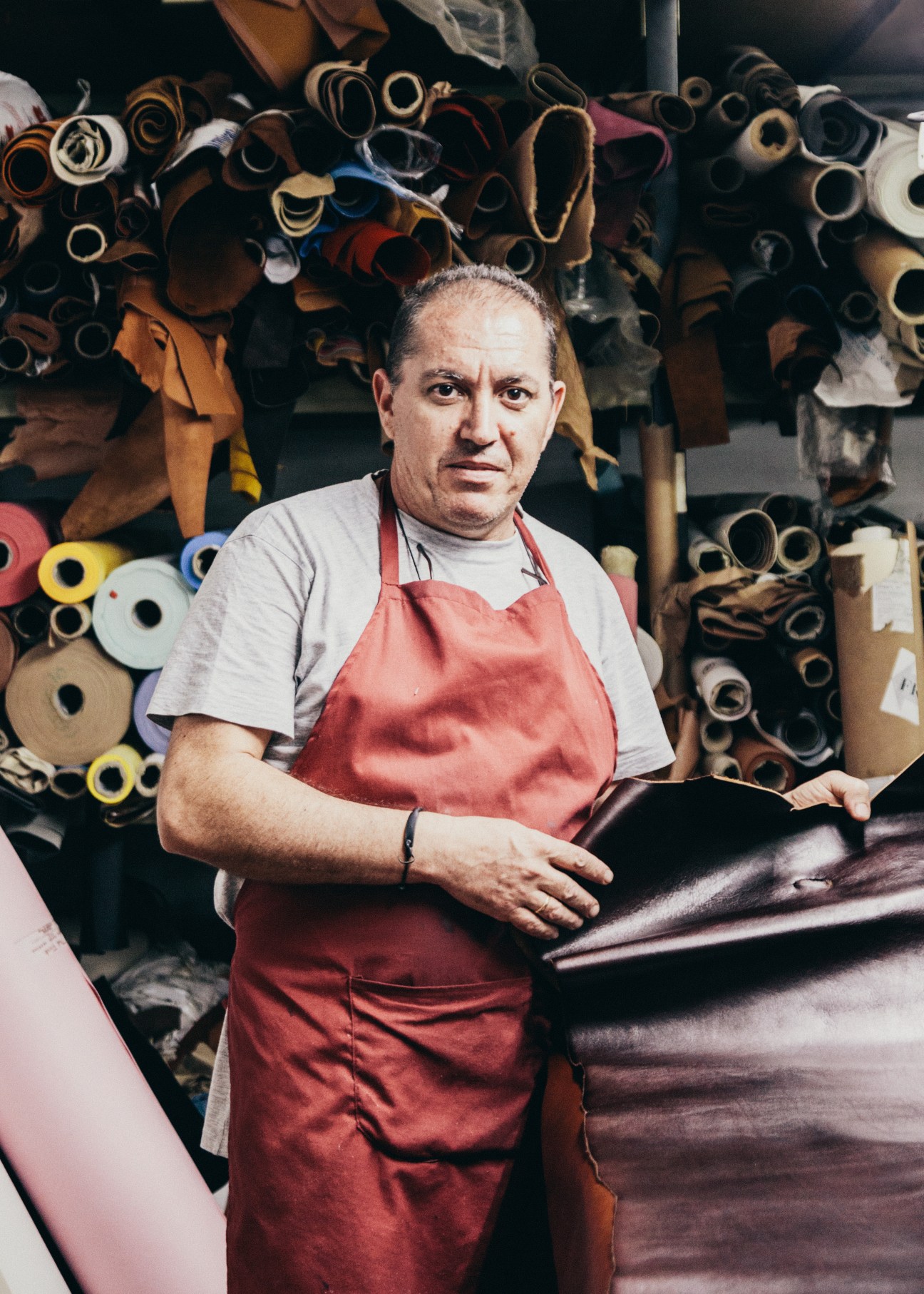 A leather worker wearing a red apron holds a leather hide in front of rolled leather swatches.
