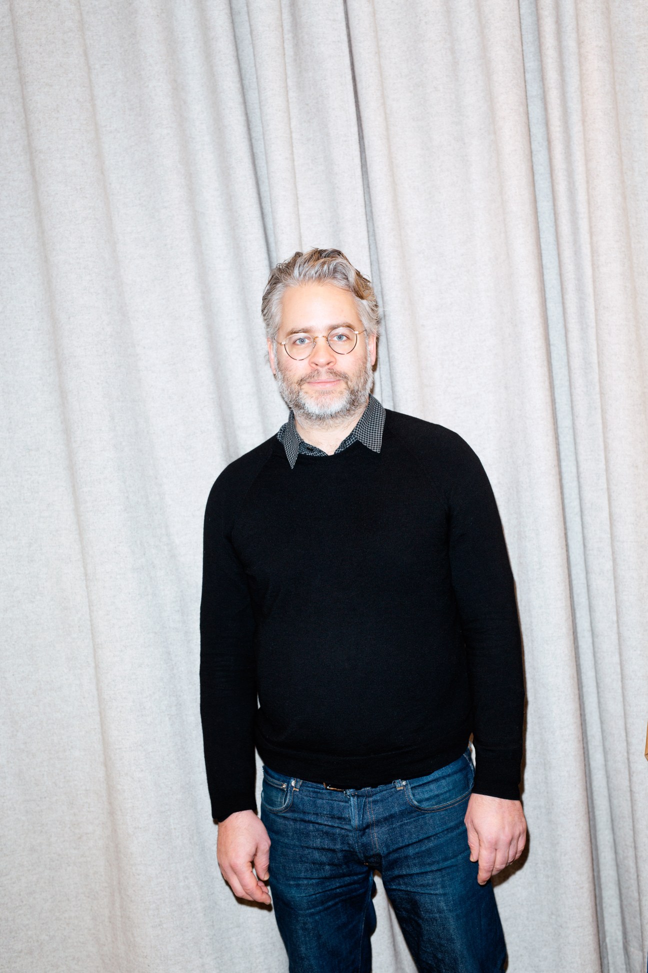 Man wearing round glasses and black sweater over checkered shirt against white curtain backdrop.