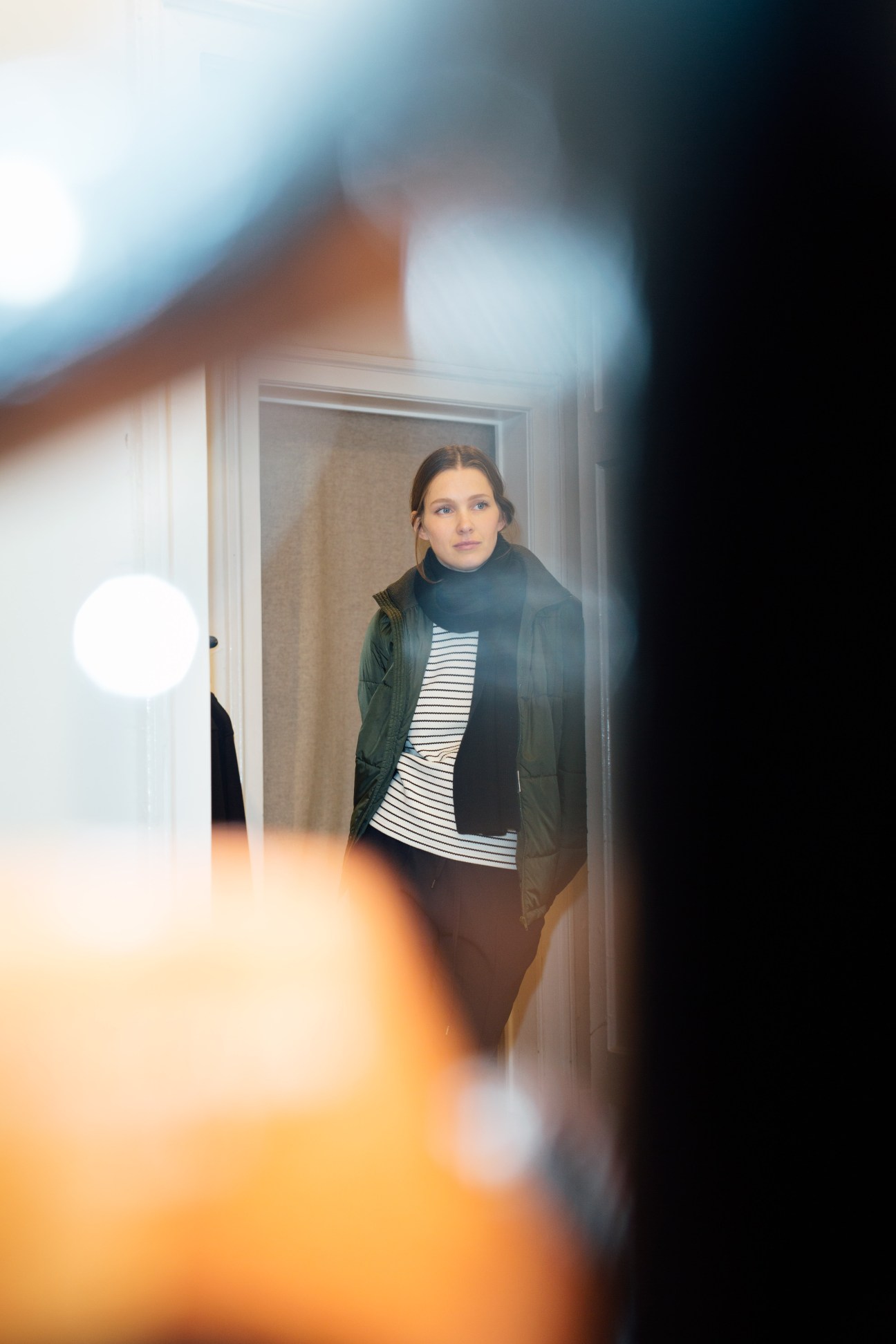 Susi Streich stands in a doorway wearing a striped shirt, black scarf, and green quilted jacket.