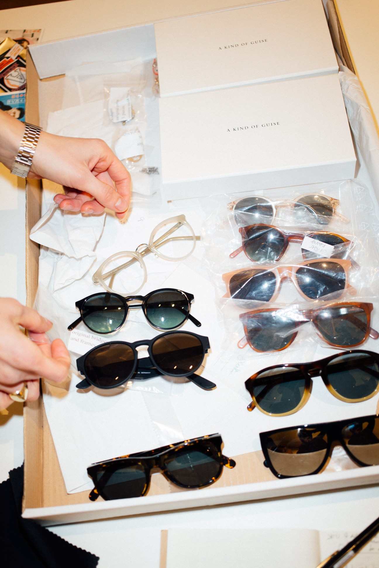 A hand holding a pair of eyeglasses above a collection of sunglasses displayed on white paper with "A Kind of Guise" branding.