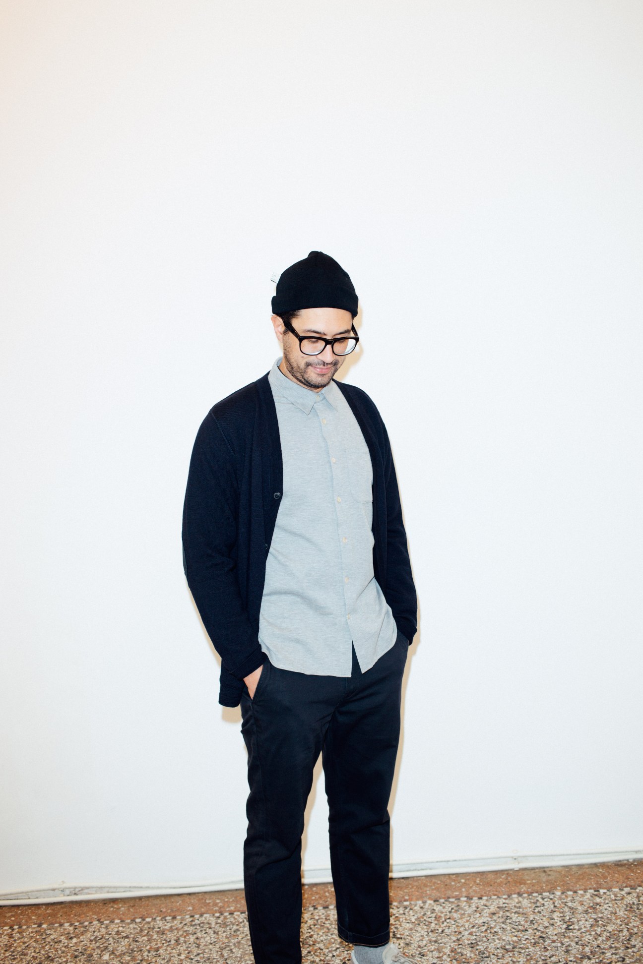 Man wearing black beanie, glasses, light blue shirt and dark cardigan standing against white background.