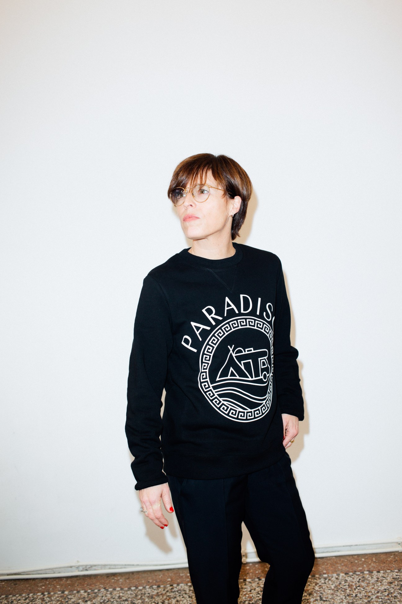 Woman wearing black Paradise sweatshirt with circular Greek key logo design, standing against white background.