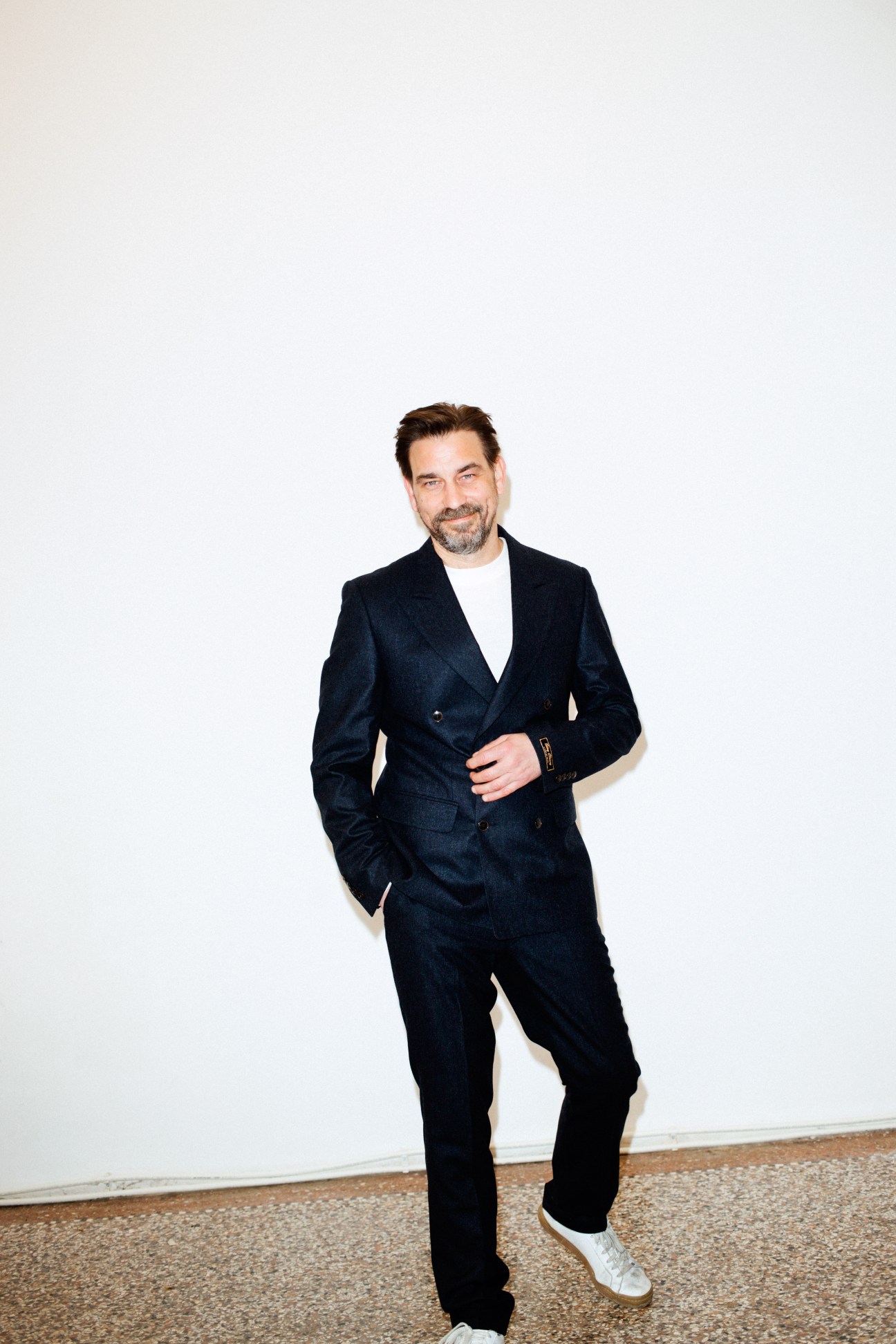 Man in dark double-breasted blazer and white sneakers standing against white wall