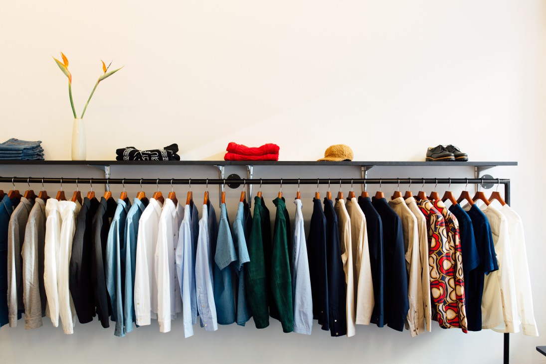 A Kind of Guise clothing rack displaying colorful garments with accessories and flowers on shelf above.