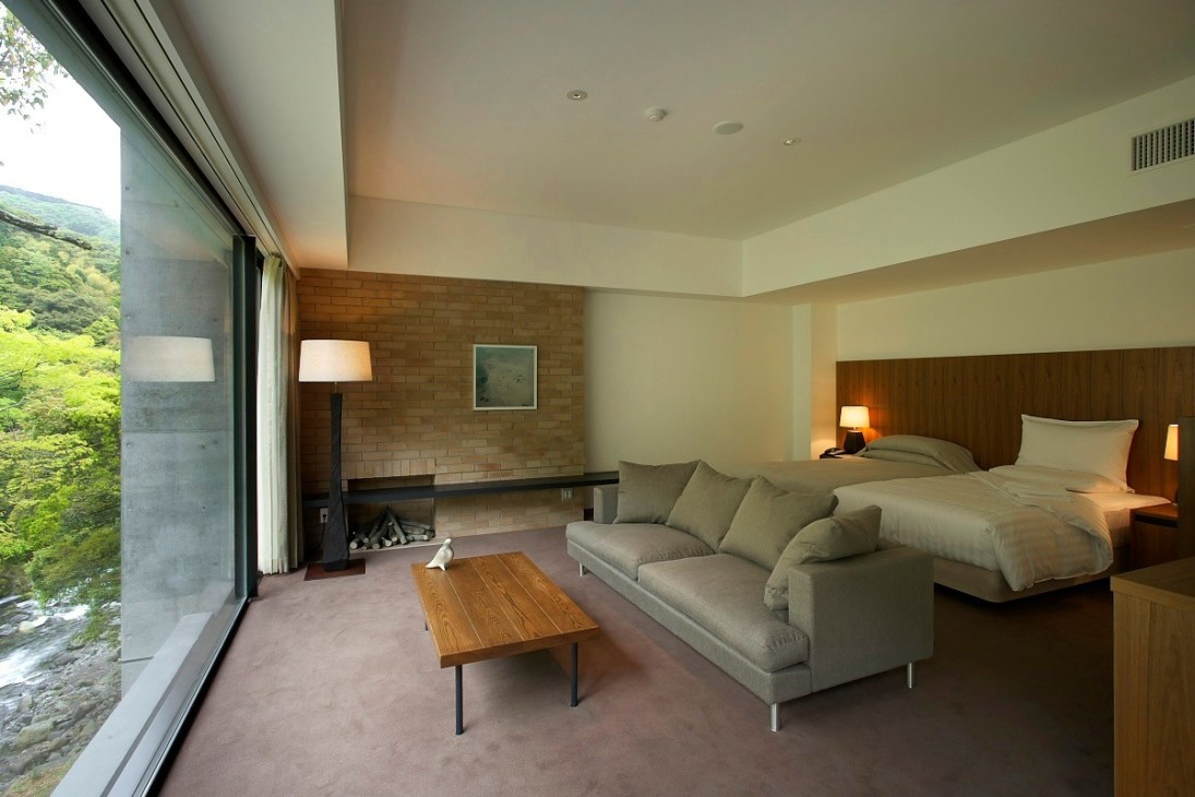 Modern hotel room at Arcana Izu with wooden furnishings, floor-to-ceiling windows overlooking greenery, and a fireplace.