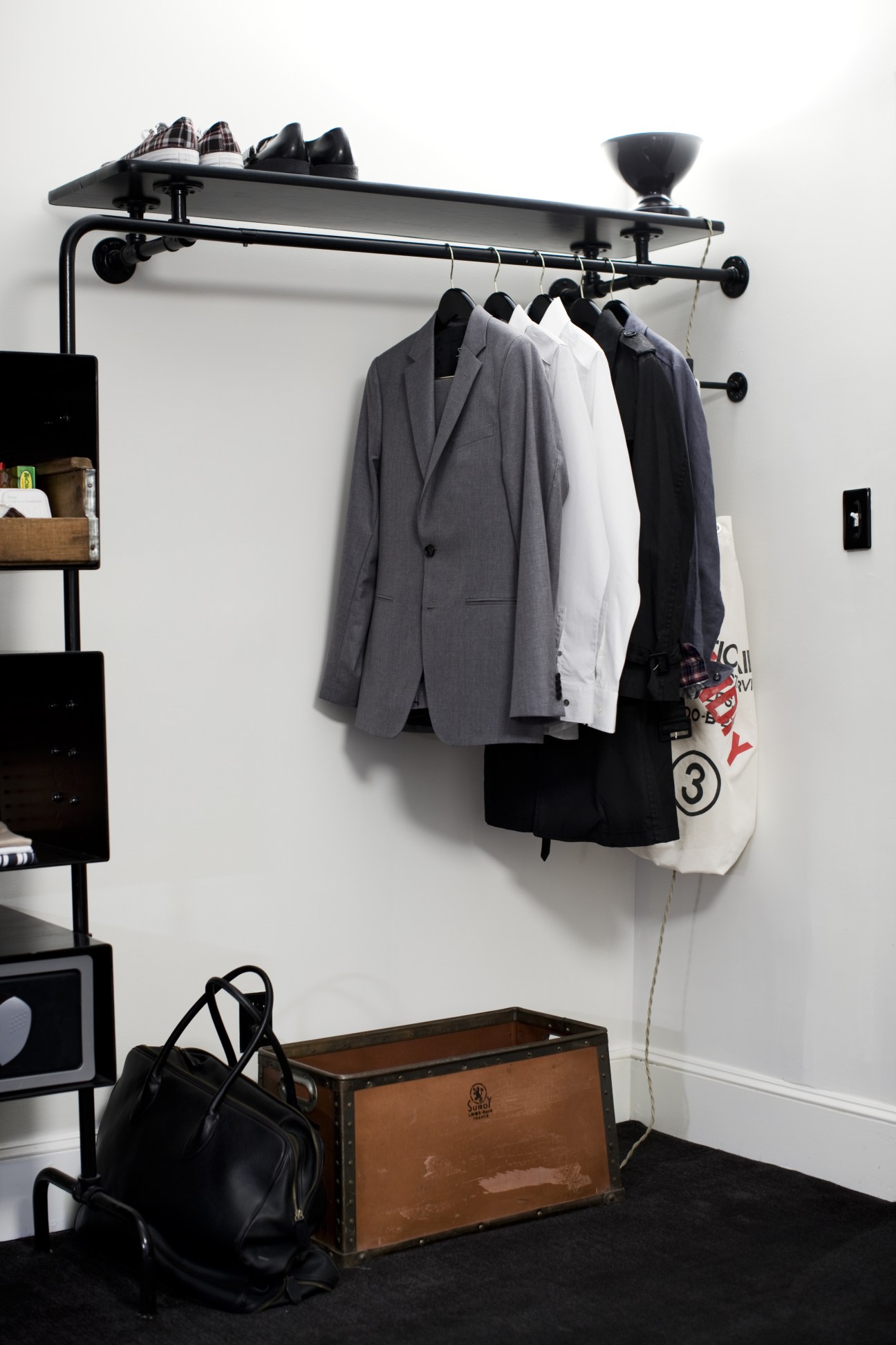 Black metal wall-mounted clothing rack with hanging jackets and blazers above a vintage brown leather trunk.