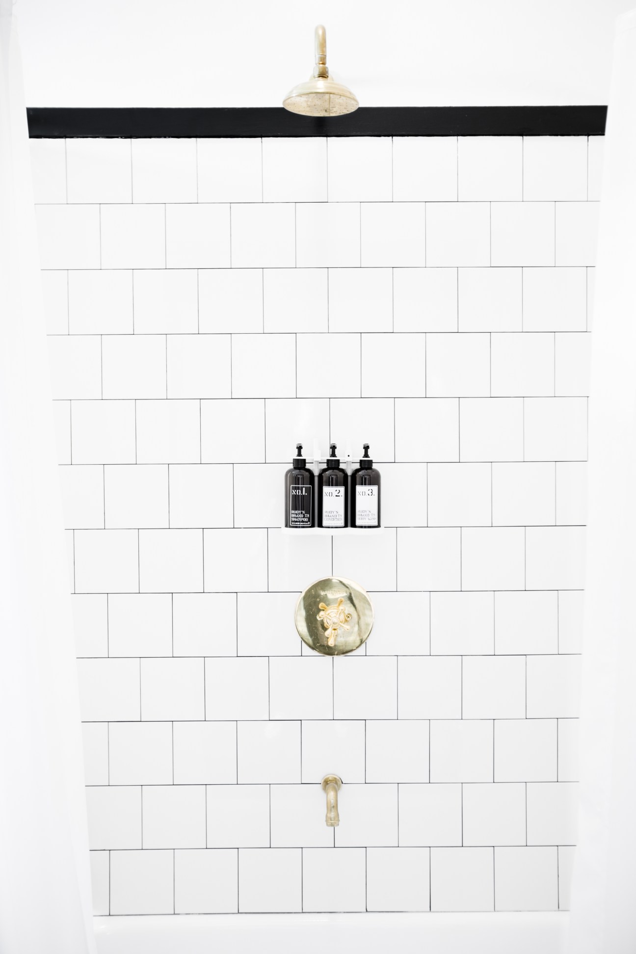 Minimalist bathroom wall with white subway tiles, brass fixtures, and three dark bottles on a shelf.