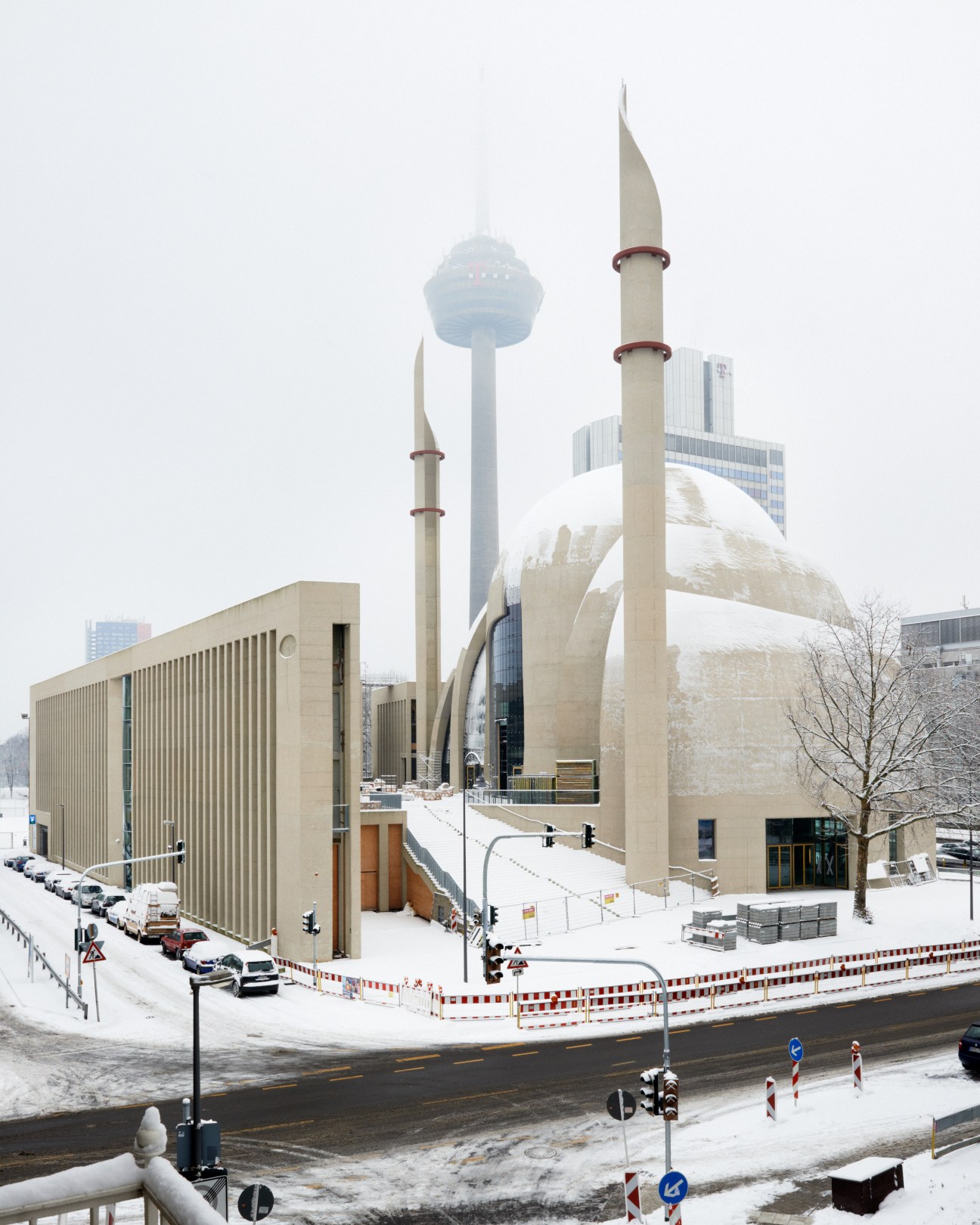 The Central Mosque in Köln's Ehrenfeld district, featuring pale concrete structures, two minarets, and a large dome covered in snow.