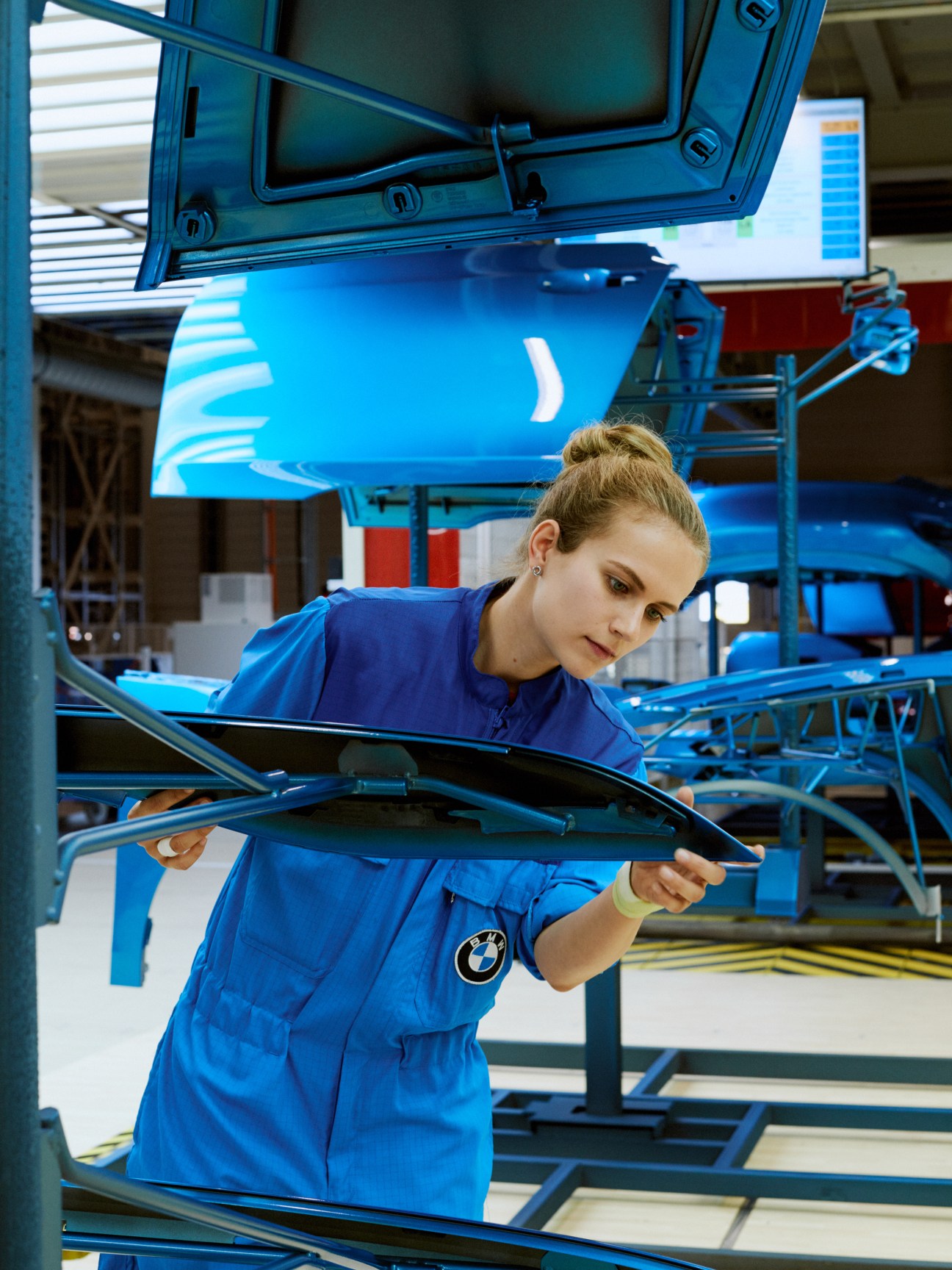 Woman in blue BMW factory uniform inspecting car part under bright light.