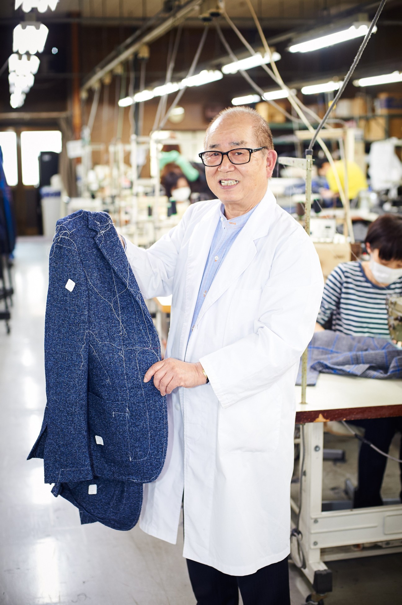 Man in white lab coat holding blue jacket on factory floor with sewing equipment in background.