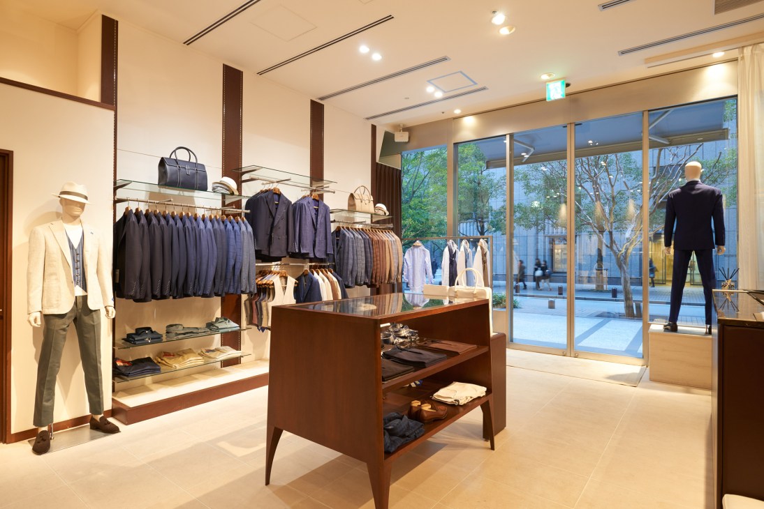 Modern menswear boutique interior with mannequins displaying suits and blazers, wooden fixtures, and large windows overlooking a street.