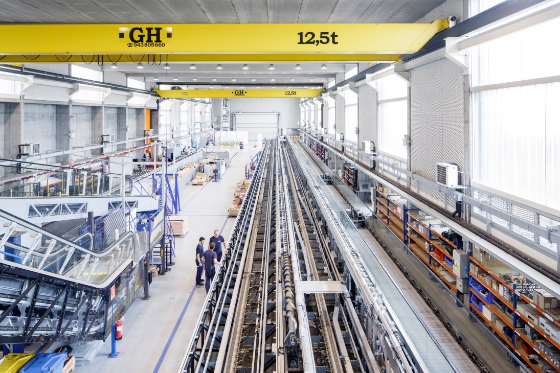 Industrial testing facility with yellow overhead cranes, rail tracks, and workers in ThyssenKrupp Elevator's innovation centre.