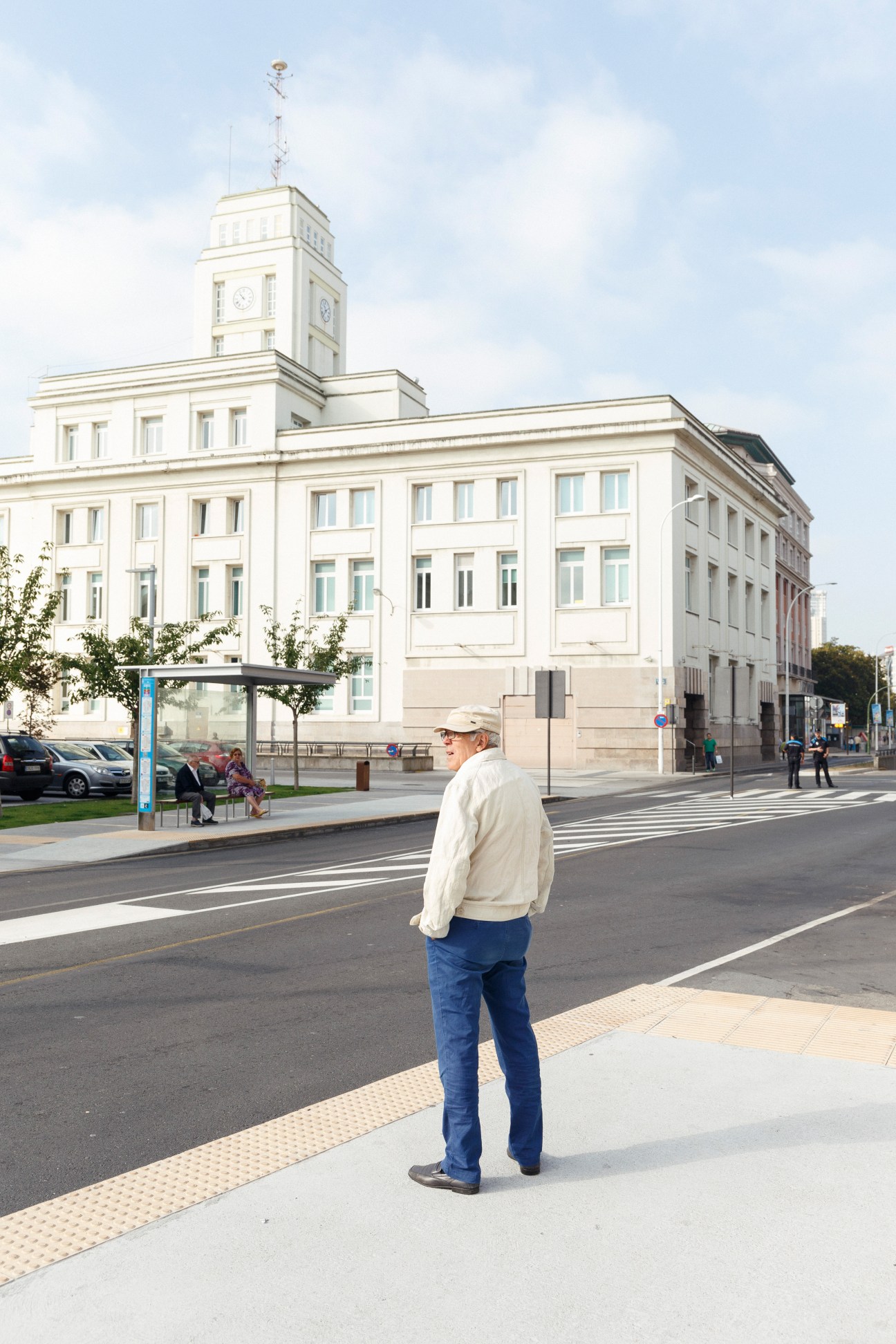 Man in light jacket and cap waiting on sidewalk in front of white neoclassical building with clock tower.
