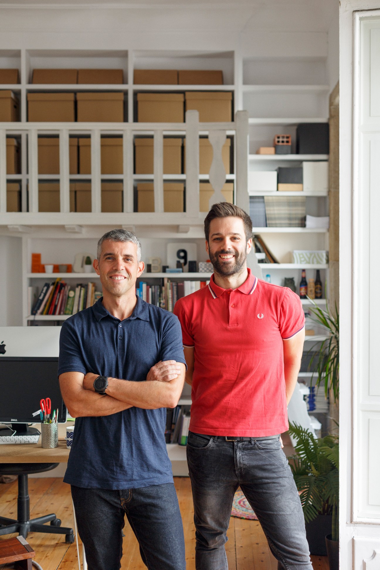 Two men standing in a modern design studio with white shelving and wooden desk.