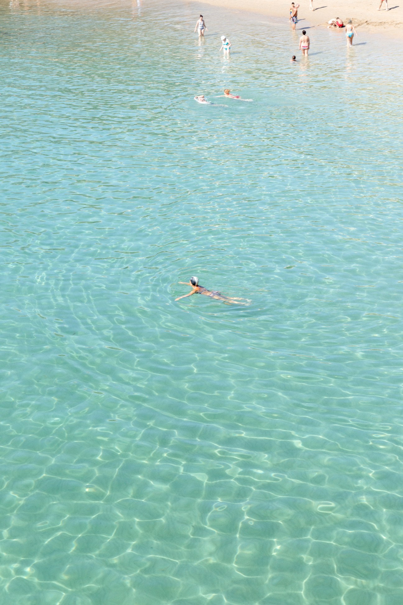 A person swimming in clear turquoise Atlantic waters with other beachgoers visible in the background.