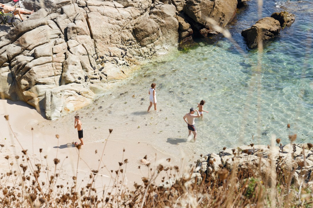 Children swimming in clear turquoise water at a rocky beach cove in A Coruña's Adormideras neighbourhood.