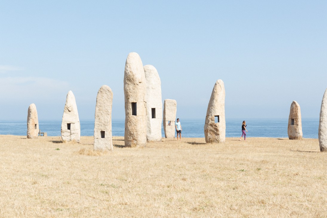 Large white granite sculptures resembling menhirs with window openings stand on a sandy beach with ocean and sky in background.