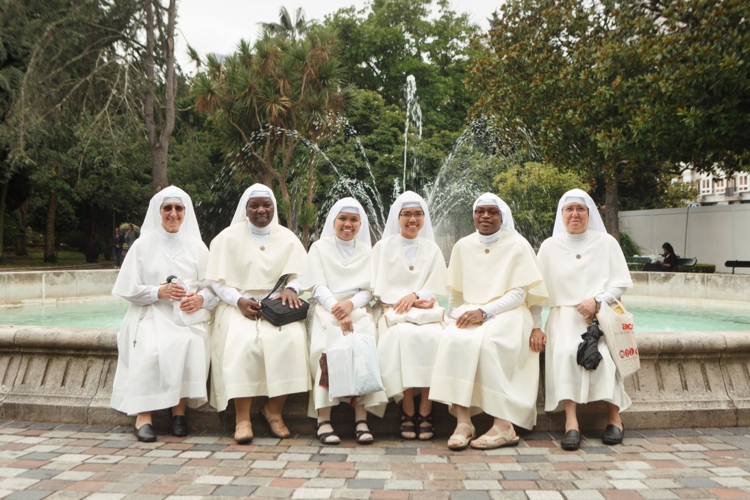 Six women in white robes and headscarves sit by a fountain in a park setting.