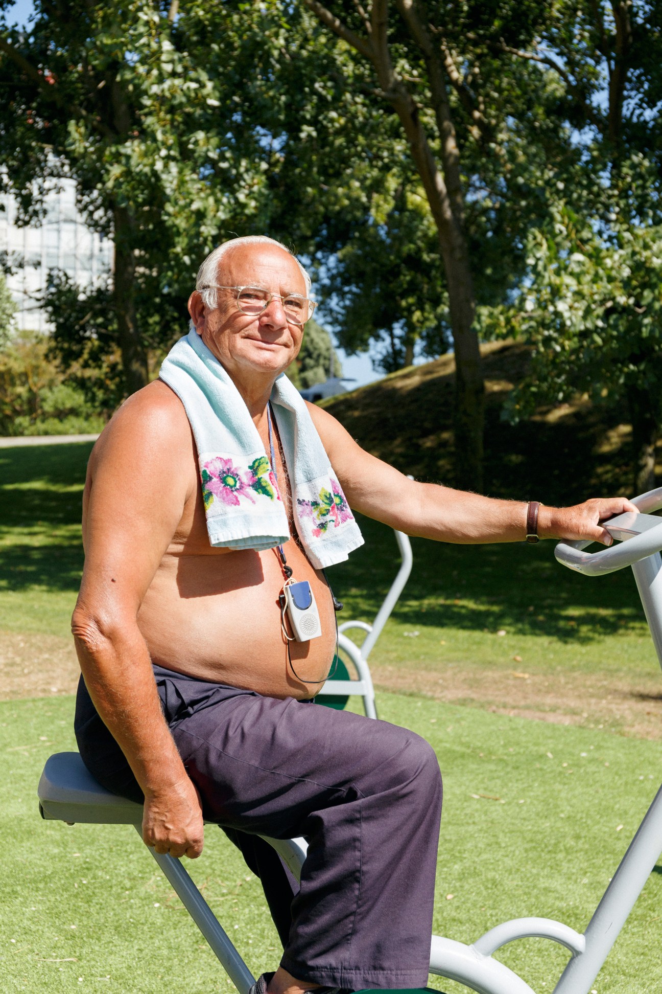 Senior man exercising on outdoor gym equipment in a park, wearing glasses and a decorative towel.