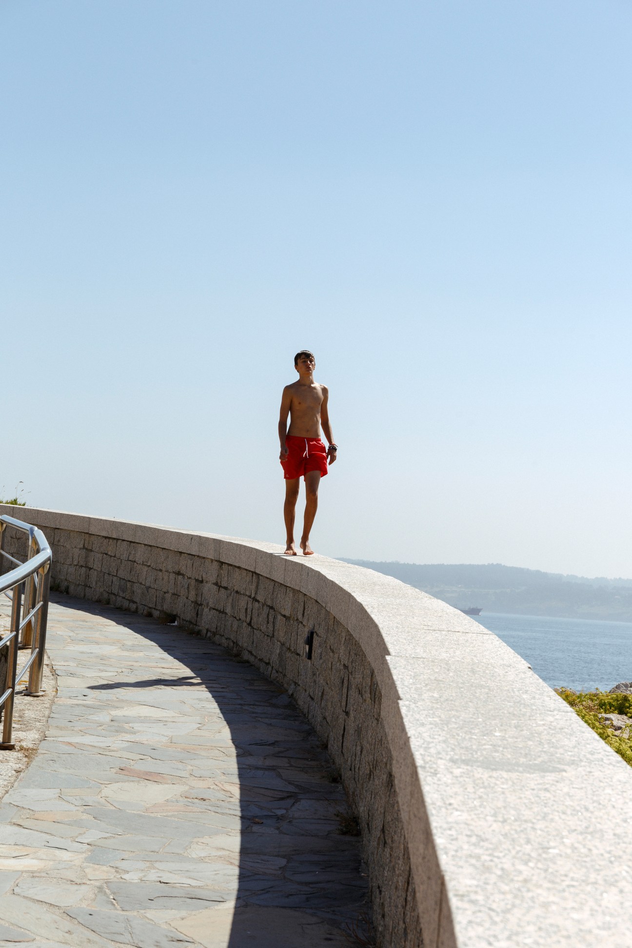 Shirtless man in red swim shorts standing on curved stone waterfront structure overlooking Atlantic Ocean.