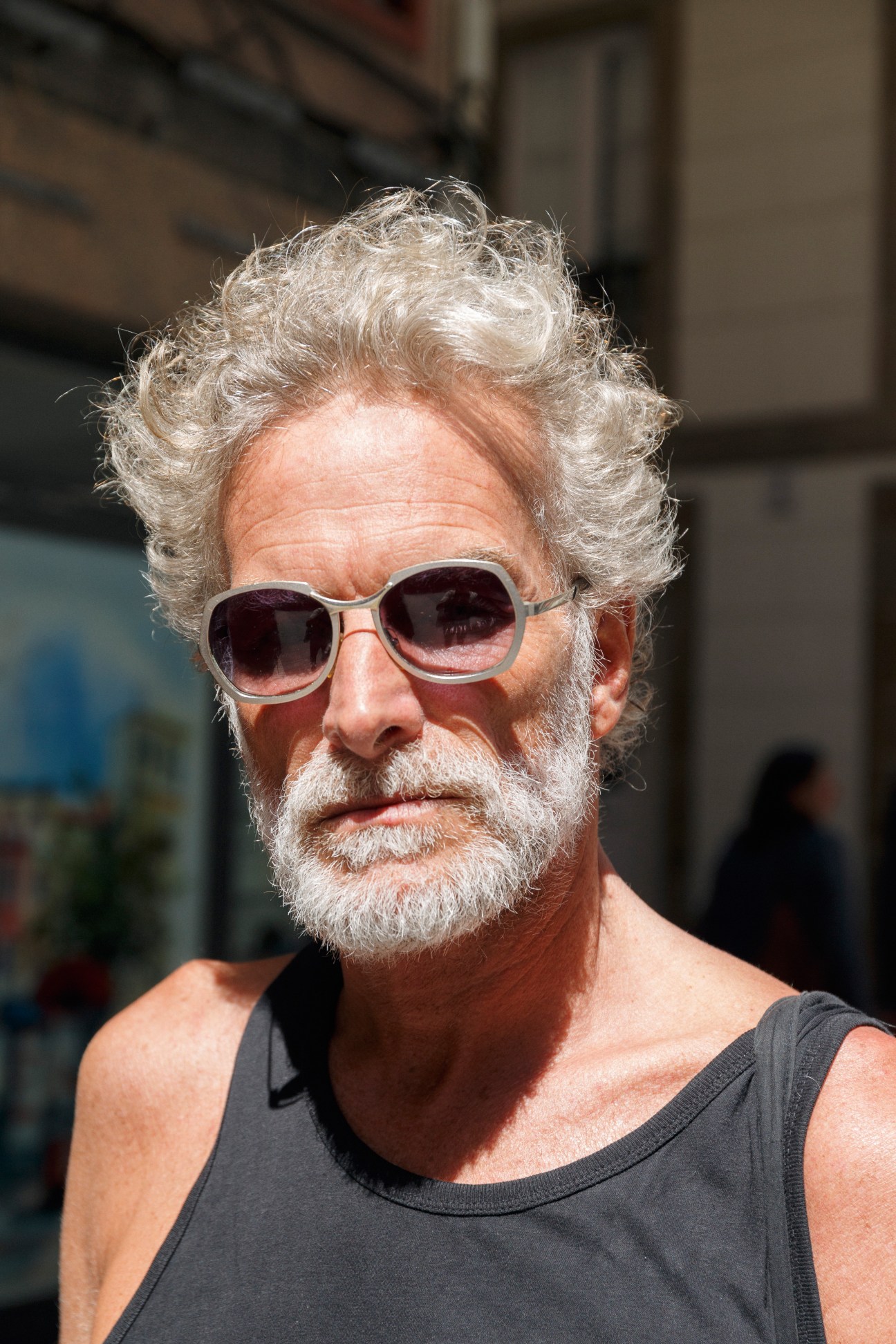Older man with white curly hair and beard wearing pink-tinted sunglasses and dark sleeveless shirt