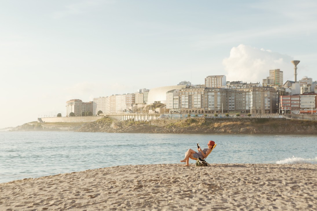 Person in red hat sitting in beach chair on sandy shore with A Coruña waterfront and buildings in background