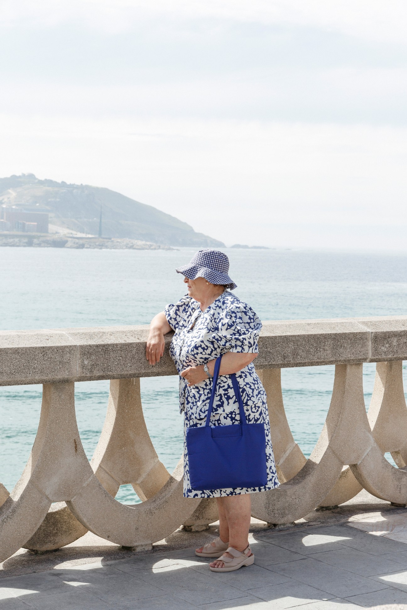 Woman in blue and white patterned dress and hat resting against stone railing on A Coruña's waterfront promenade with ocean and headland in background.
